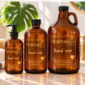 PurposeFill hand soap in 16 oz, 32 oz and 64 oz reusable amber glass bottles with gold lettering, displayed beside a bathroom sink with monstera greenery.